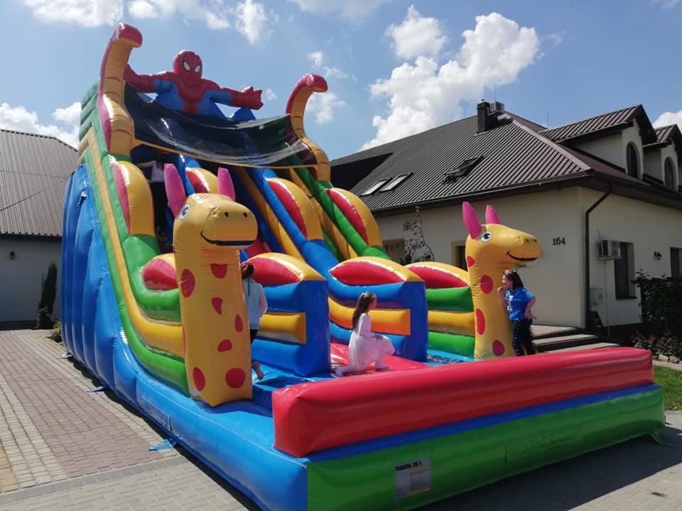 Dmuchana zjeżdżalnia z motywem Spider-Mana i żyraf, ustawiona na brukowanym podwórku przed domem z numerem 164, dzieci bawiące się na konstrukcji.