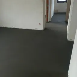 Świeżo wylana, ciemnoszara posadzka w surowym wnętrzu budynku z widocznymi fragmentami ścian z cegły i oknem w tle.