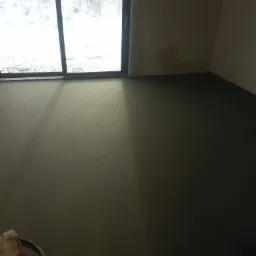 Świeżo wylana, gładka posadzka jastrychowa w pomieszczeniu z dużym oknem balkonowym, przez które widać zimowy krajobraz.