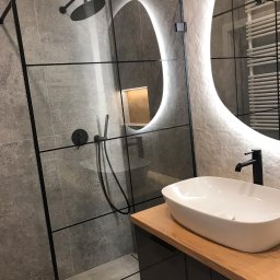Dekorroom - Nowoczesna łazienka z kabiną prysznicową typu walk-in z czarnymi profilami, owalną umywalką nablatową na drewnianym blacie i podświetlanym lustrem o nieregularnym kształcie.