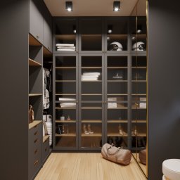 Dekorroom - Nowoczesna garderoba z szarymi szafami, przeszklonymi drzwiami i drewnianymi półkami, oświetlona punktowo, z torebką podróżną na podłodze i dekoracyjną figurką na parapecie.