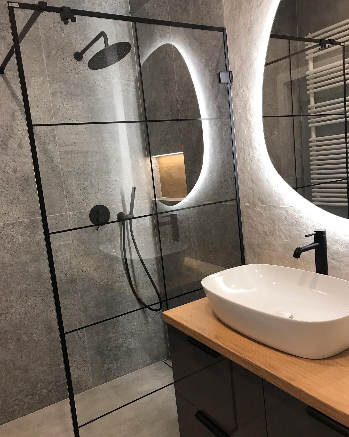 Nowoczesna łazienka z kabiną prysznicową typu walk-in z czarnymi profilami, owalną umywalką nablatową na drewnianym blacie i podświetlanym lustrem o nieregularnym kształcie.