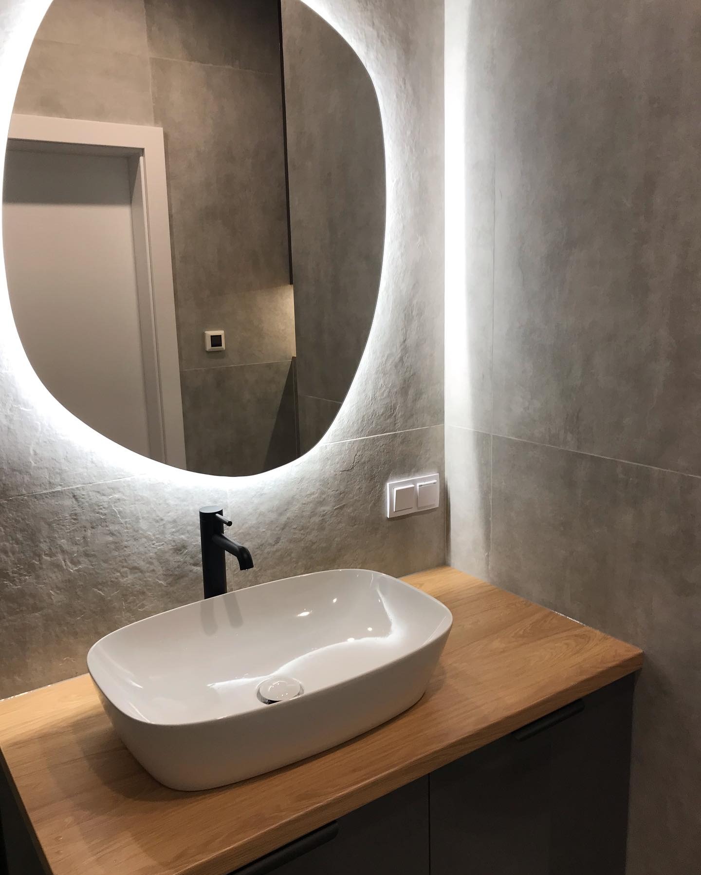 Nowoczesna łazienka z owalnym lustrem LED, białą umywalką nablatową i czarną armaturą, wykończona płytkami w odcieniach szarości, z drewnianym blatem pod umywalką.