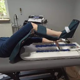 Pacjentka leżąca na stole rehabilitacyjnym, poddana terapii z wykorzystaniem urządzenia do ćwiczeń stawu kolanowego, widoczny panel sterowania i podłączenia do nóg.