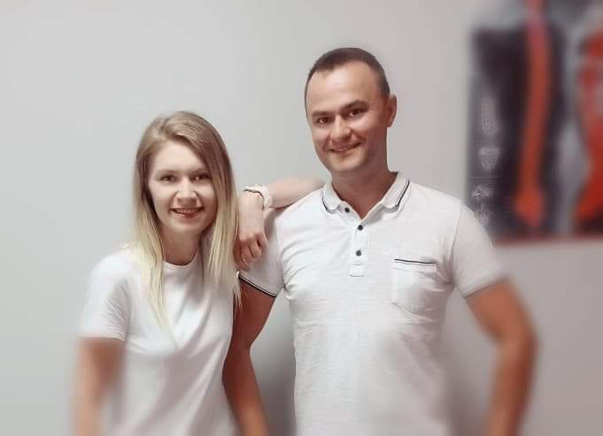 Uśmiechnięta kobieta i mężczyzna w białych koszulkach, pozujący w gabinecie z plakatem anatomicznym w tle.