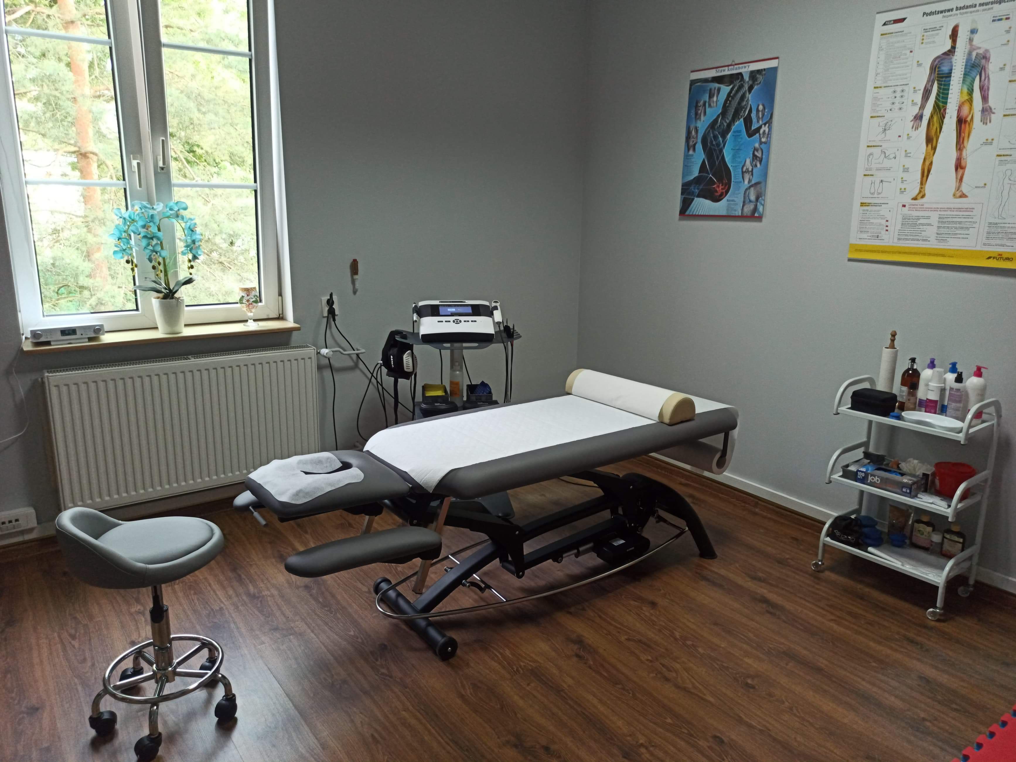Gabinet z profesjonalnym stołem do zabiegów, taboretem na kółkach, aparaturą do fizykoterapii i wózkiem z akcesoriami, doświetlony naturalnym światłem z okna.