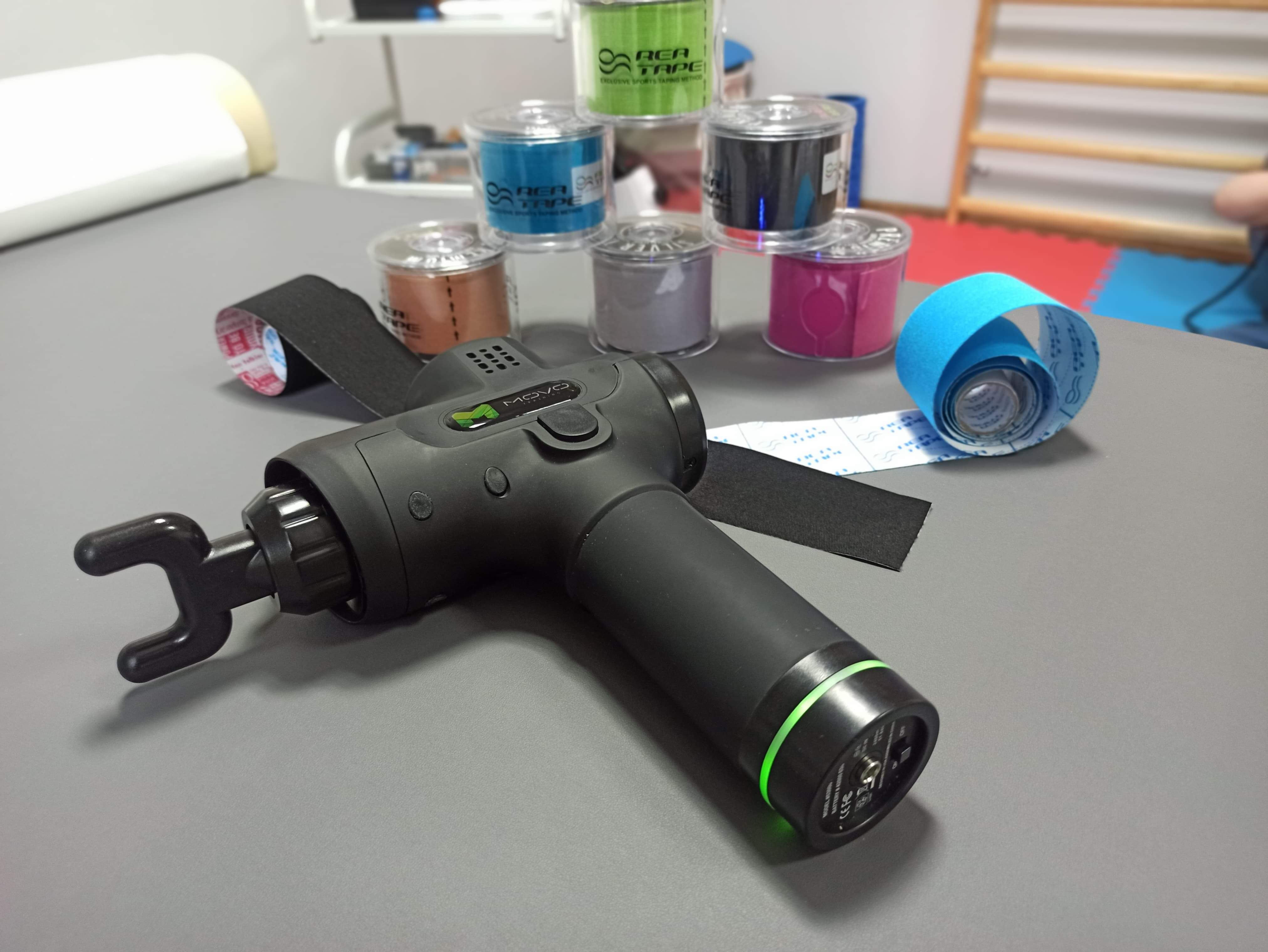 Pistolet do masażu MOVO leżący na szarym stole, obok kolorowych rolek taśmy do kinesiotapingu marki REA TAPE, z fragmentem drabinki gimnastycznej w tle.