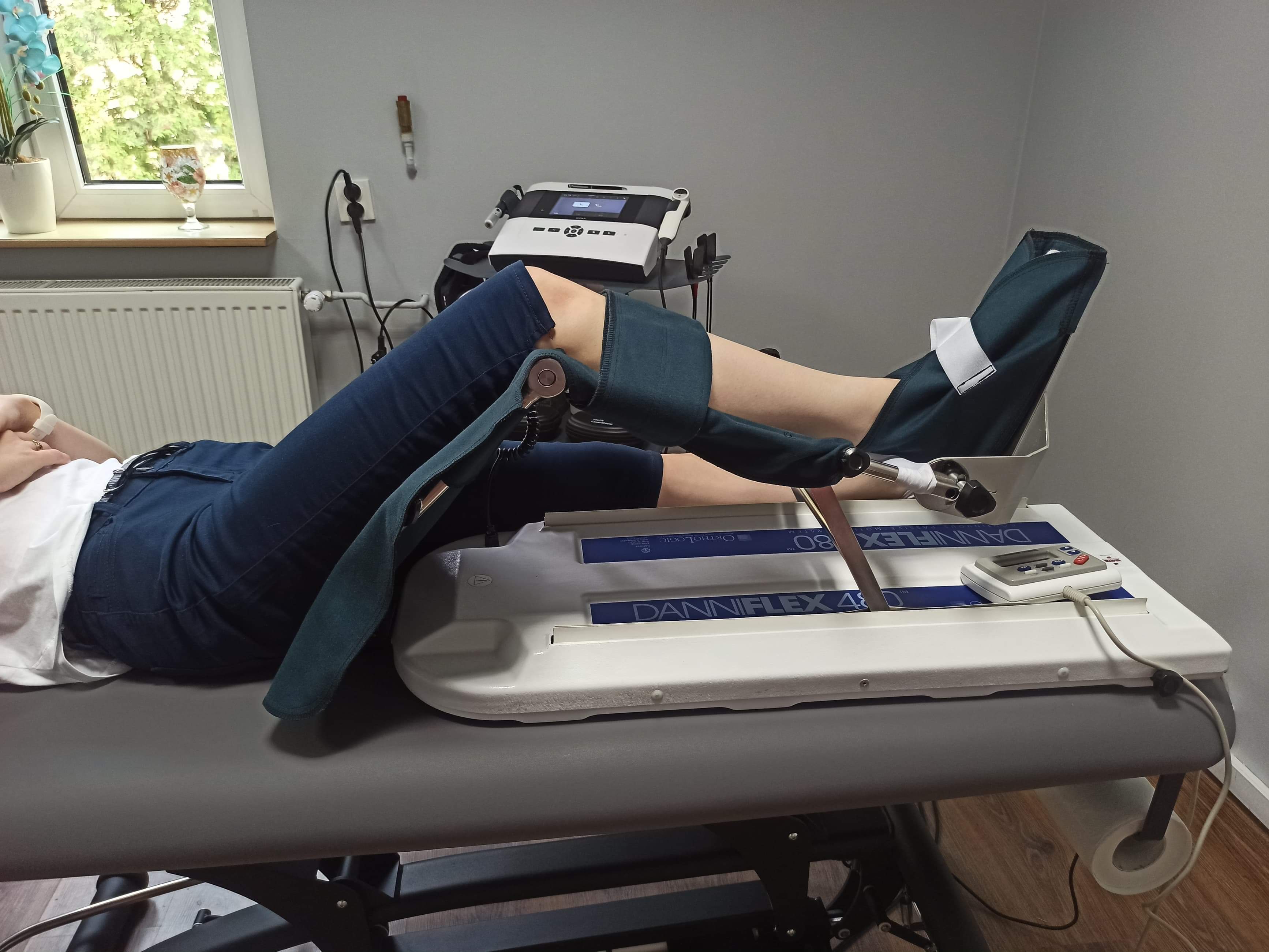 Pacjentka leżąca na stole rehabilitacyjnym, poddana terapii z wykorzystaniem urządzenia do ćwiczeń stawu kolanowego, widoczny panel sterowania i podłączenia do nóg.