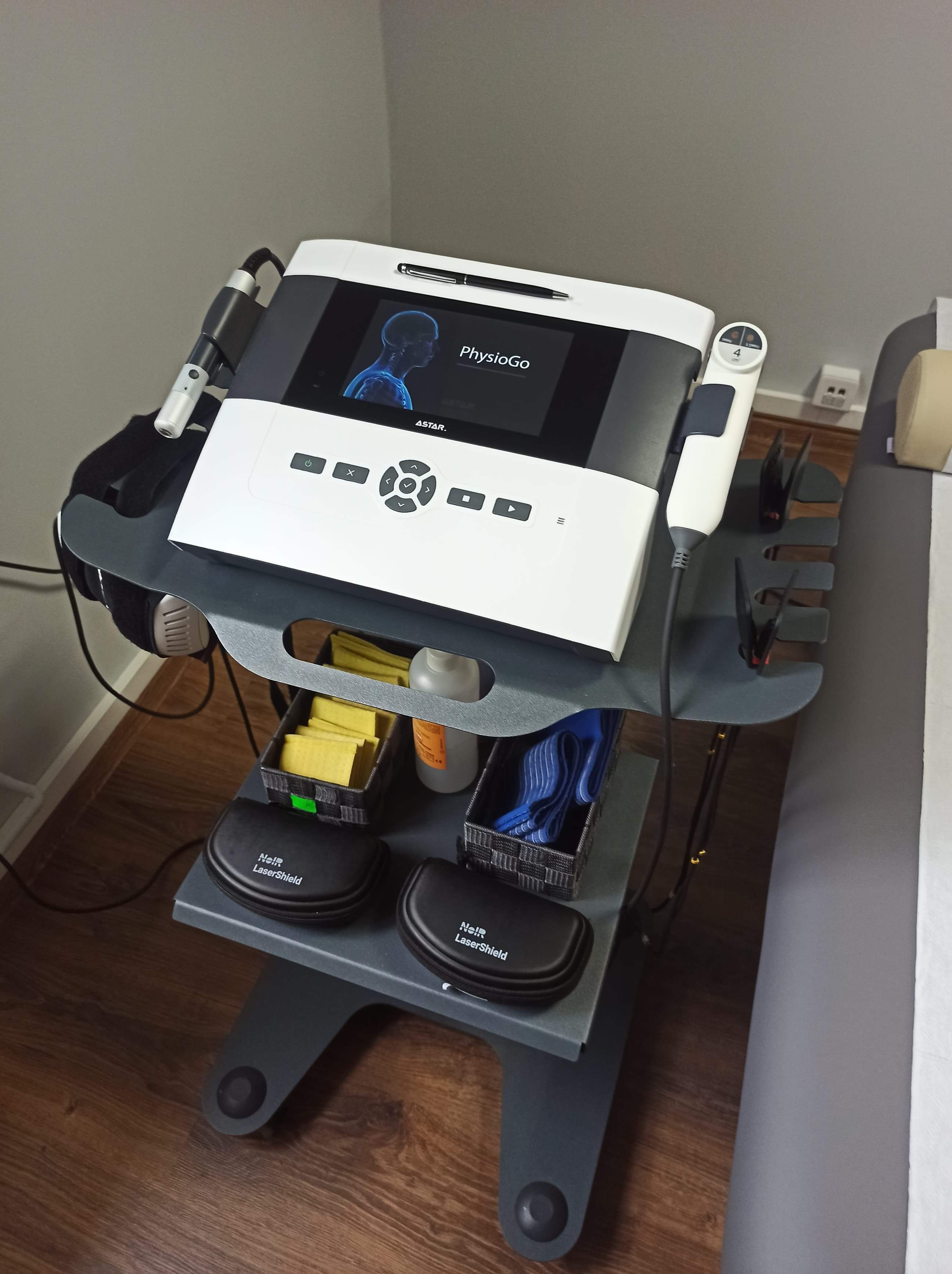 Profesjonalny sprzęt do fizykoterapii PhysioGo Astar na wózku z akcesoriami, w tym aplikatorami, żelami i osłonami laserowymi Noir LaserShield, ustawiony w gabinecie.