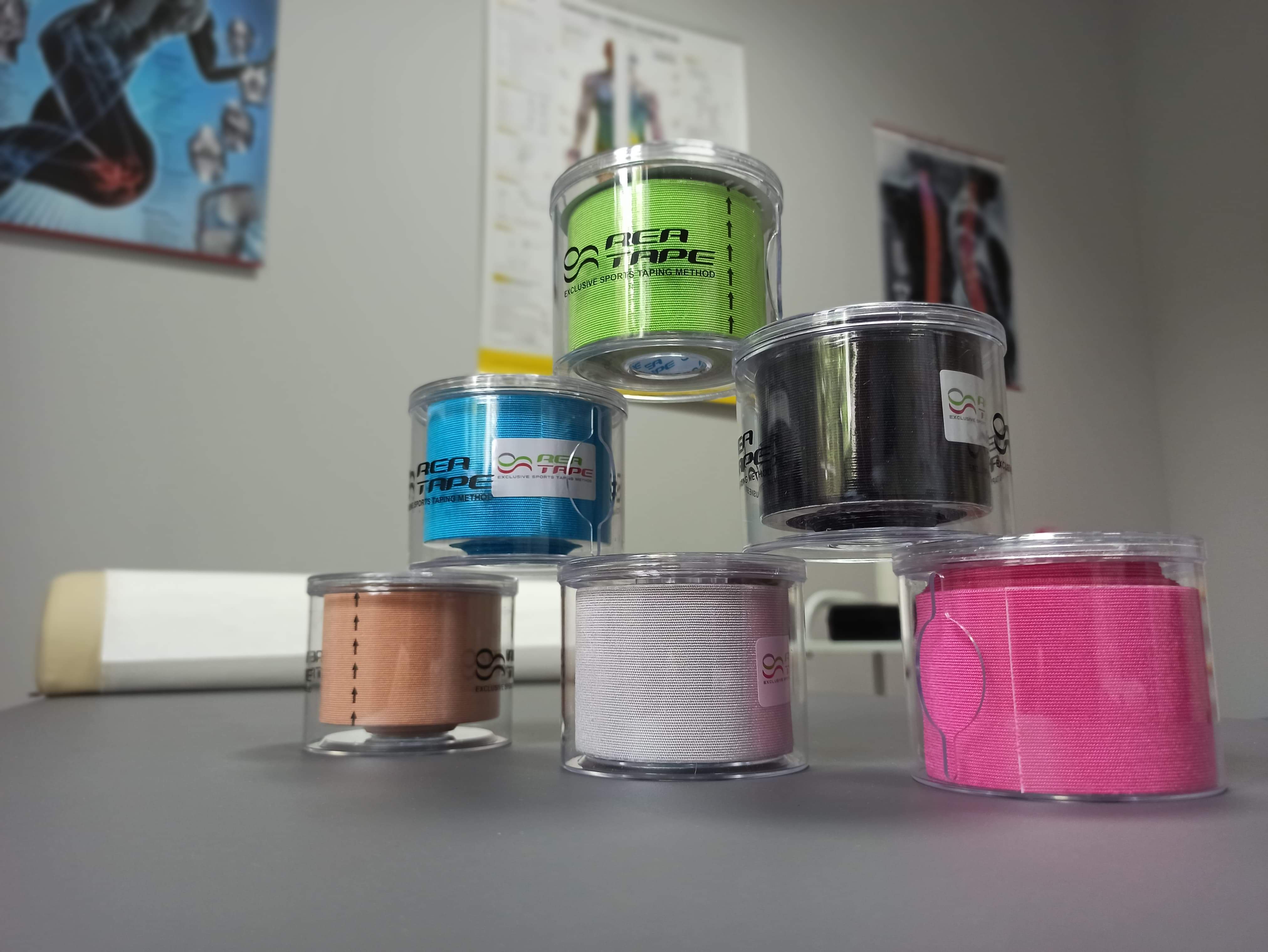 Piramida z kolorowych rolek elastycznych taśm kinezjologicznych w plastikowych opakowaniach, widoczne plakaty anatomiczne w tle.