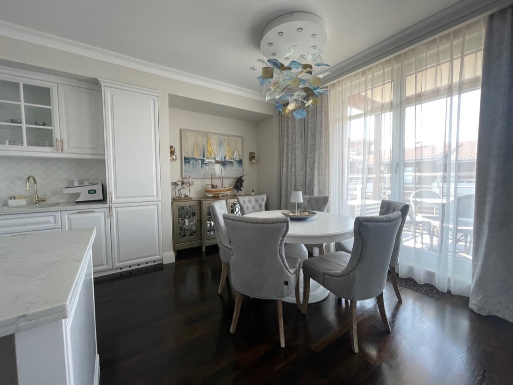 Jasna jadalnia w stylu nadmorskim: biały okrągły stół otoczony szarymi krzesłami, dekoracyjna lampa z motywem morskim, widok na balkon przez przeszklone drzwi.