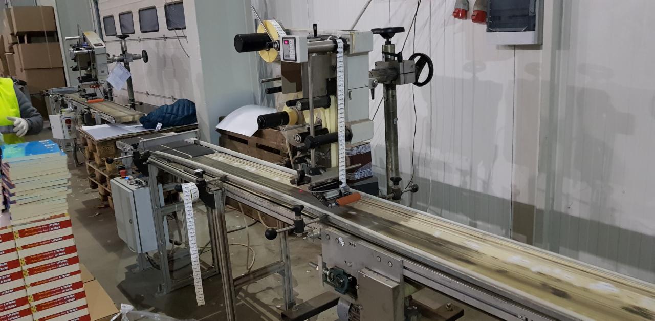 Automatyczna linia etykietująca w hali produkcyjnej, z widocznymi książkami na palecie i rolką etykiet.