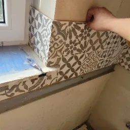 Układanie dekoracyjnych płytek ceramicznych wzorzystych przy oknie w trakcie prac wykończeniowych, widoczny fragment okna, wiadro z klejem i szczotka.