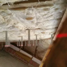 Ocieplenie poddasza wełną mineralną, widoczne metalowe wieszaki i czerwona linia lasera poziomującego.