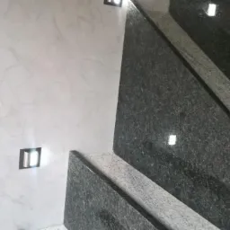 Ściana z dekoracyjną, perłową strukturą w odcieniach szarości i białe, kwadratowe oprawy oświetleniowe, sąsiadująca z ciemnymi, granitowymi schodami z delikatnym połyskiem.