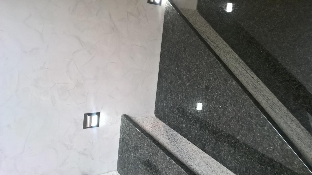 Ściana z dekoracyjną, perłową strukturą w odcieniach szarości i białe, kwadratowe oprawy oświetleniowe, sąsiadująca z ciemnymi, granitowymi schodami z delikatnym połyskiem.