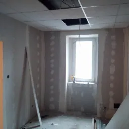 Wnętrze w trakcie remontu: ściany z widocznymi łączeniami płyt, sufit podwieszany z otworami, okno z narzędziami na parapecie, płyty gotowe do montażu na podłodze.