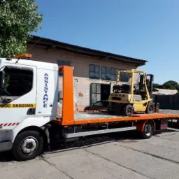 Żółty wózek widłowy Toyota transportowany na lawecie pomocy drogowej z napisem Assistance i Pomoc Drogowa 24h na tle budynku.