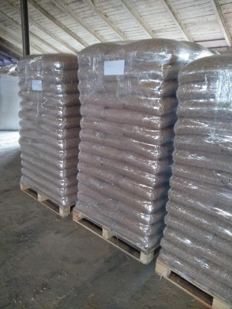 Palety z workami pelletu owinięte folią stretch, składowane w magazynie z drewnianą konstrukcją dachu.