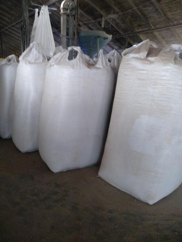 Rząd białych worków typu Big Bag wypełnionych pelletem drzewnym w magazynie, widoczna instalacja do napełniania worków w tle, podłoga lekko zabrudzona.