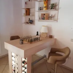 Drewniany blat z szachami i oświetleniem LED, wsparty na konstrukcji z półkami na wino, uzupełniony o krzesło barowe i dekoracyjne półki ścienne.