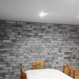 Ściana akcentowa z szarej cegły w jadalni, oświetlona dwoma punktowymi lampami sufitowymi, z widocznym fragmentem drewnianego stołu i krzesłami.