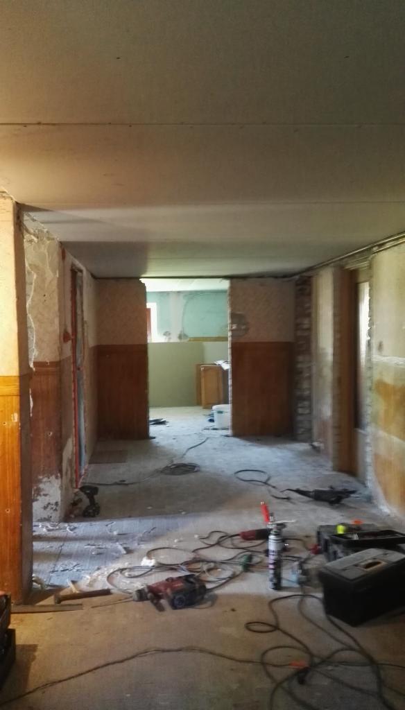 Wnętrze w trakcie demontażu, widoczne ślady po tapetach, odsłonięte fragmenty ścian, narzędzia budowlane na podłodze, w tle widoczny fragment innego pomieszczenia.