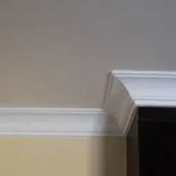 Białe listwy dekoracyjne na styku sufitu i ściany w jasnym wnętrzu, widoczne fragmenty ciemnych mebli.