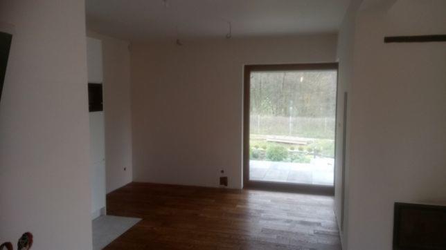 Wnętrze pokoju z drewnianą podłogą w trakcie prac wykończeniowych, ściany przygotowane do malowania, widoczne gniazdko elektryczne i duże okno z widokiem na ogród.