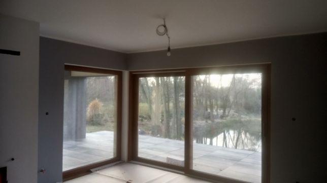 Surowe wnętrze z dużymi oknami z widokiem na jezioro i las, szare ściany, widoczny kabel od oświetlenia sufitowego, przygotowane do dalszych prac wykończeniowych.
