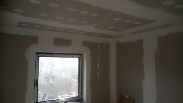 Pokój w trakcie remontu: ściany i sufit z płyt kartonowo-gipsowych, widoczne łączenia przed szpachlowaniem, sufit podwieszany z otworami, przewód zwisający z sufitu, okno z widokiem na zewnątrz.
