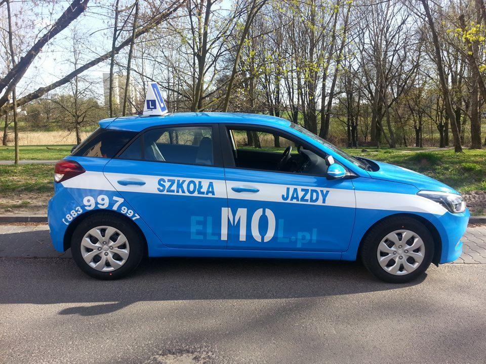 Niebieski samochód oznaczony napisem 'Szkoła Jazdy' i symbolem 'L' na dachu, zaparkowany na ulicy w słoneczny dzień, z widocznym parkiem w tle.