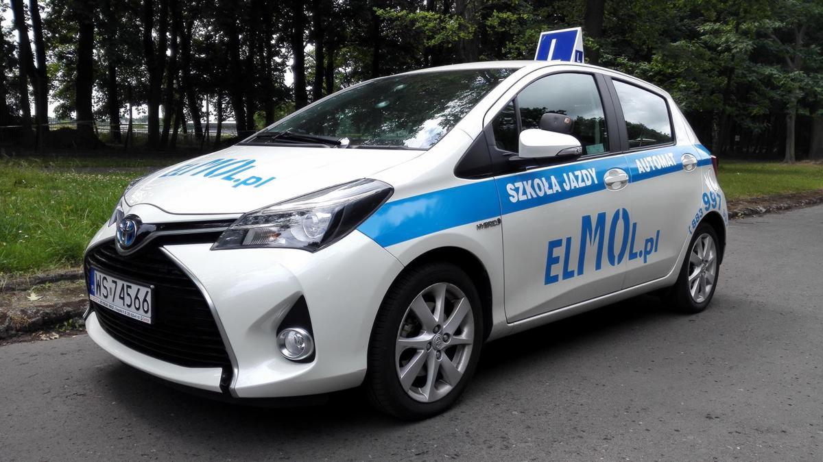 Biały samochód Toyota Yaris z niebieskim pasem i napisem 'Szkoła Jazdy' oraz 'ELMOL.pl' na tle zieleni drzew, widoczny znak 'L' na dachu.