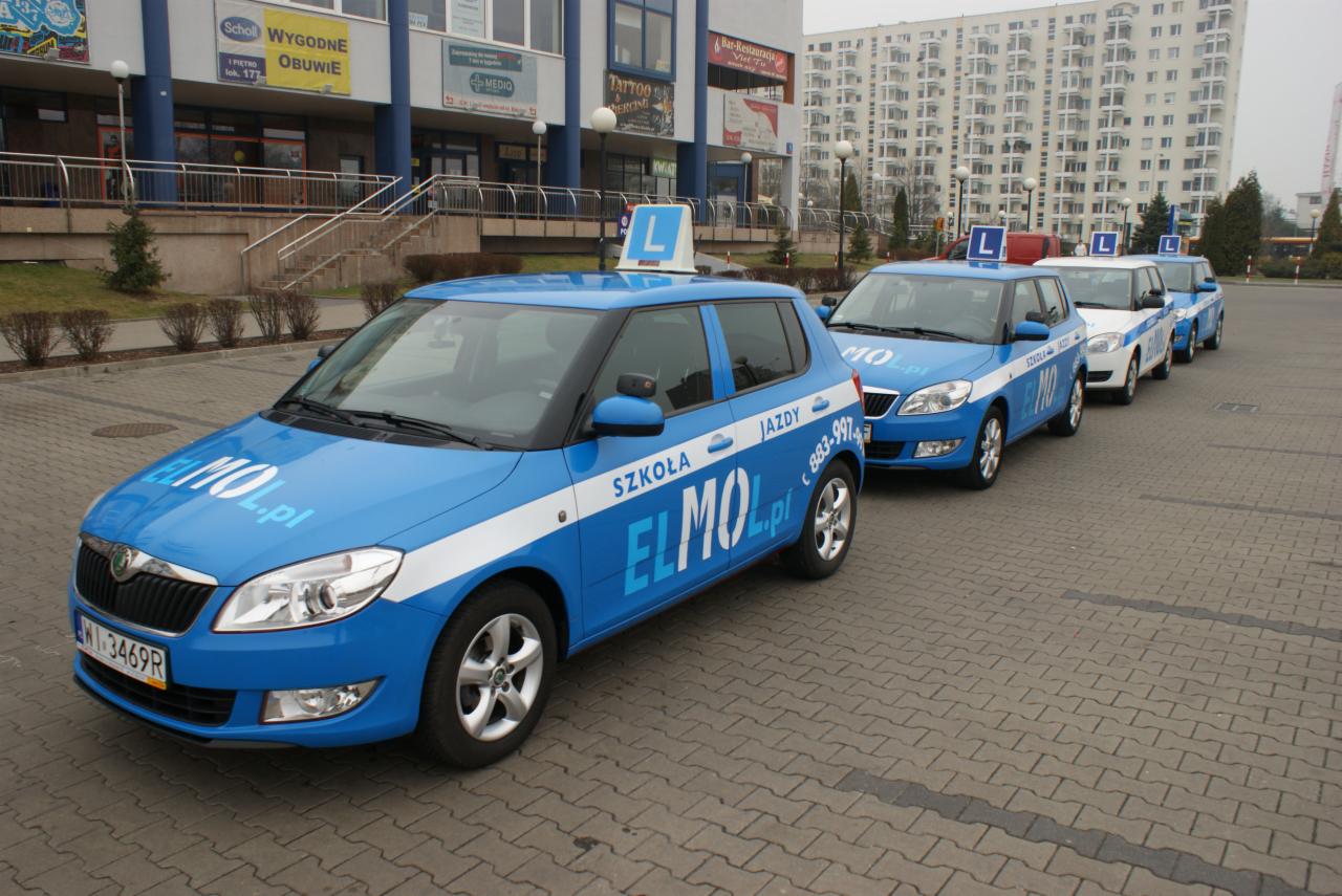 Flota niebieskich samochodów z oznaczeniami szkoły nauki jazdy ustawiona rzędem na parkingu przed budynkiem.