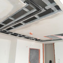 BART-HAUS - Sufit podwieszany w trakcie montażu: metalowa konstrukcja, płyty gipsowe, instalacja elektryczna. Widoczne zabezpieczone folią drzwi i fragment ściany.