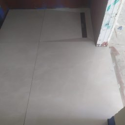 BART-HAUS - Jasne płytki podłogowe w małej łazience z odpływem liniowym, widok z góry. Ściany w trakcie wykończenia, widoczne surowe krawędzie i ślady farby.