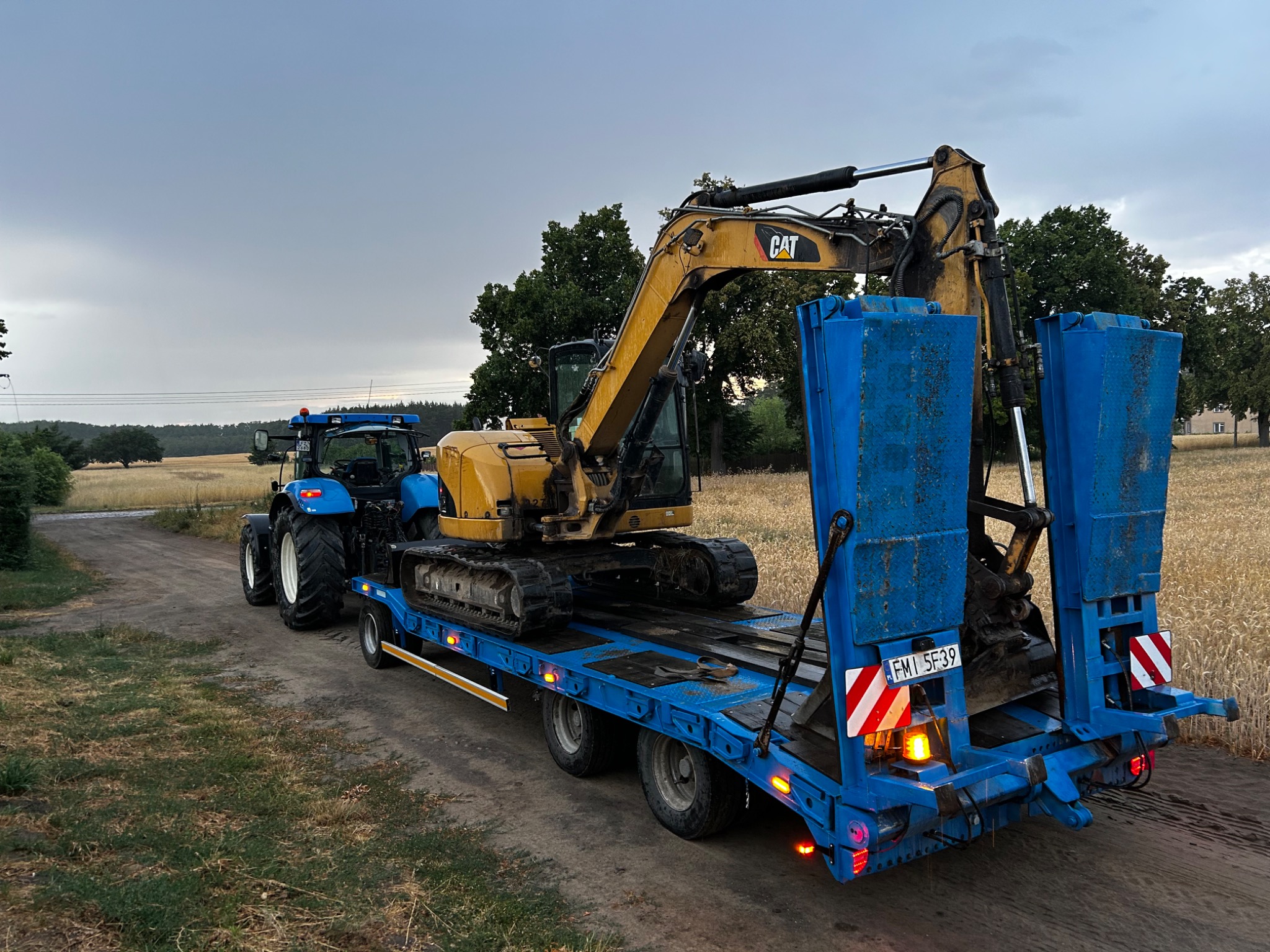 Transport koparki CAT na niskopodwoziowej lawecie ciągniętej przez niebieski traktor na tle pola i drzew. Sprzęt gotowy do pracy.