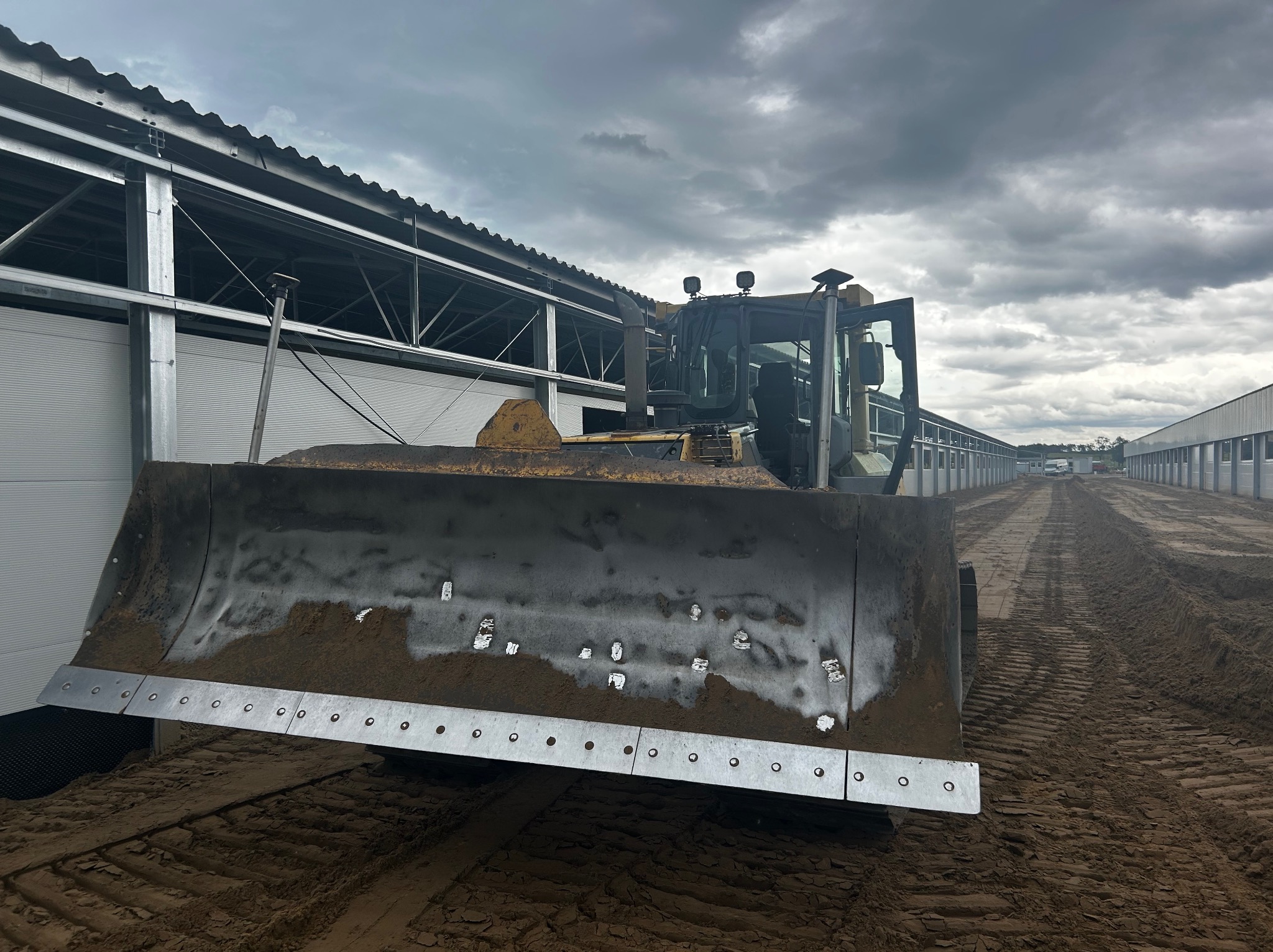 Żółta spycharka z metalowym lemieszem, na ubitej ziemi, na tle konstrukcji stalowej hali i pochmurnego nieba. Widok z przodu, lekki kąt.