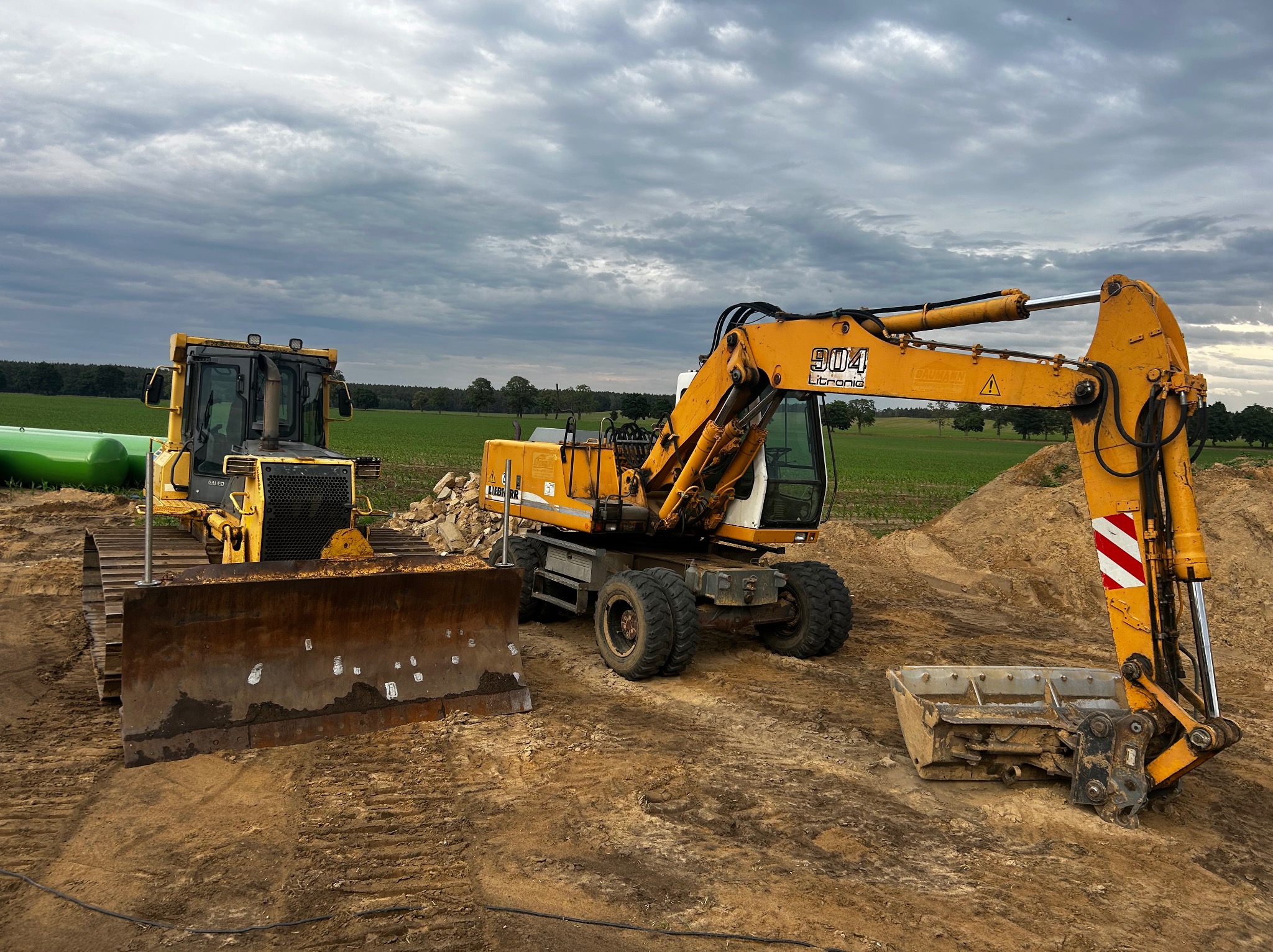 Żółty spychacz i koparka Liebherr 904 na placu budowy, z zielonymi zbiornikami w tle i pochmurnym niebem. Sprzęt ciężki gotowy do pracy.