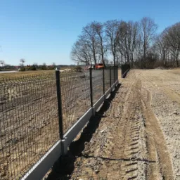 Nowo zainstalowane, czarne ogrodzenie panelowe z betonową podmurówką na nierównym terenie, z widocznymi śladami opon i budynkiem w tle.