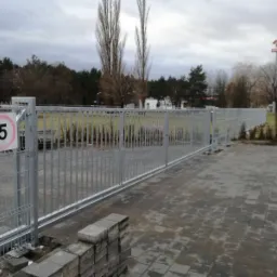 Nowo zamontowana, długa, metalowa brama przemysłowa z pionowymi prętami, widoczny znak ostrzegawczy i ograniczenie prędkości do 5 km/h, ułożona kostka brukowa, drzewa w tle, pochmurne niebo.