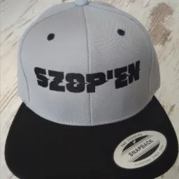 Szara czapka typu snapback z czarnym daszkiem i napisem 'SZOP'EN' wykonanym czarną czcionką, leżąca na jasnej powierzchni.