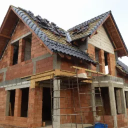 Niedokończony dom z czerwonej cegły z częściowo ułożonym, czarnym dachem z dachówki ceramicznej, widoczne rusztowania i więźba dachowa.