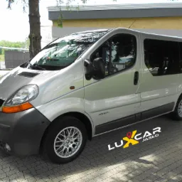 Srebrny Renault Trafic z logo Luxcar Jasło na kostce brukowej, widoczne odbicia drzew w oknach, oświetlenie uliczne w tle.