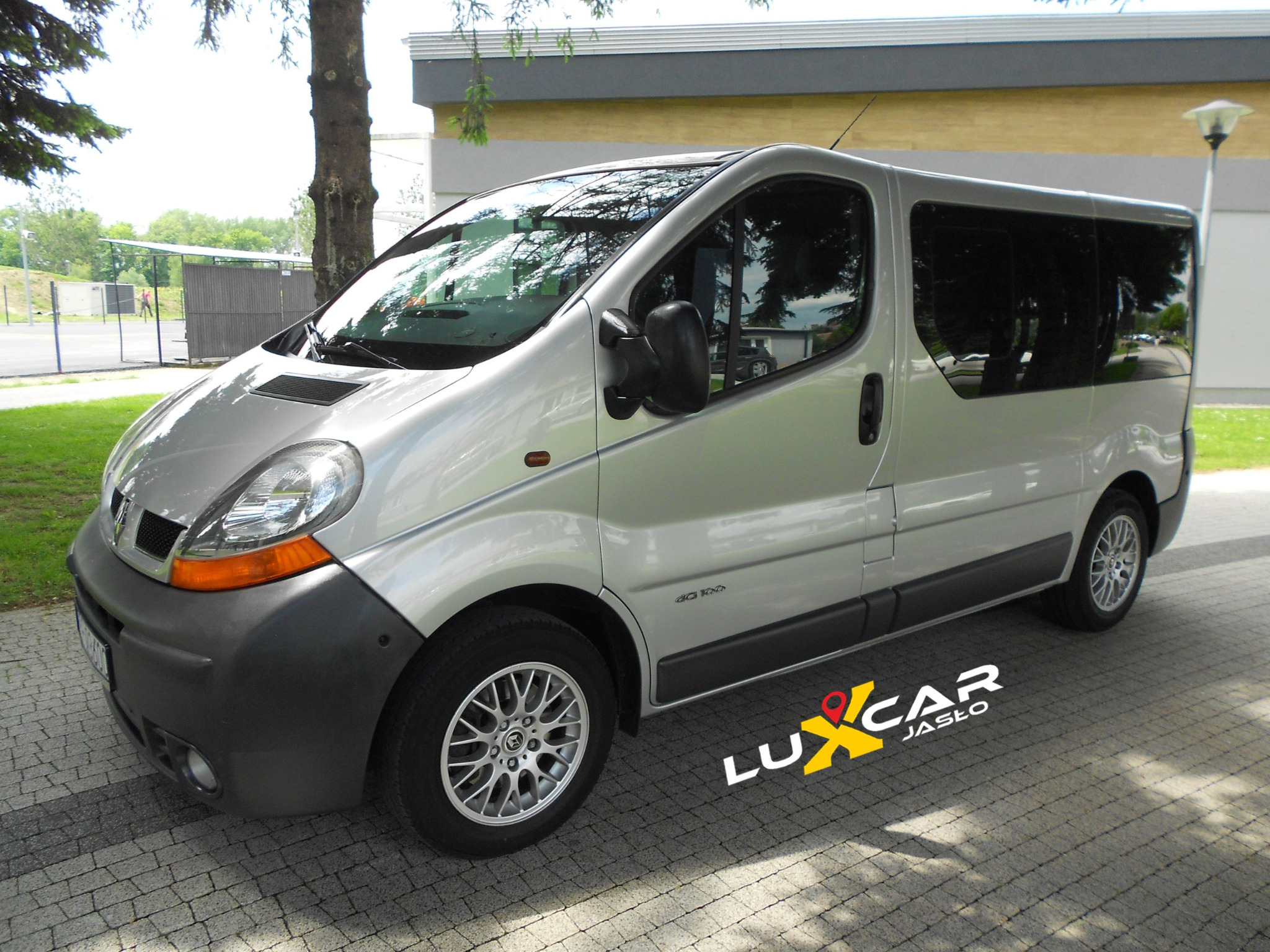 Srebrny Renault Trafic z logo Luxcar Jasło na kostce brukowej, widoczne odbicia drzew w oknach, oświetlenie uliczne w tle.