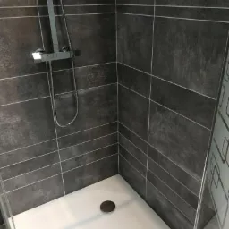 Nowoczesny prysznic z szarymi, poziomymi płytkami i białą brodzikiem z odpływem. Widoczna chromowana bateria natryskowa i fragment szklanej kabiny z wzorem.