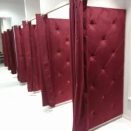 Rząd bordowych, pikowanych przymierzalni z zasłonami w sklepie odzieżowym, oświetlonych przez okrągłe lampy sufitowe.