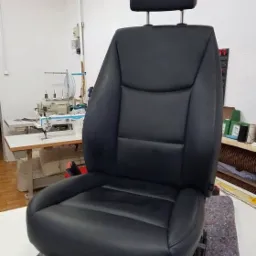Odnowione czarne skórzane siedzenie samochodowe z zagłówkiem, widoczne w warsztacie z maszyną do szycia w tle.