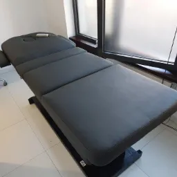 Szary, tapicerowany stół do masażu z regulowanym zagłówkiem, ustawiony w jasnym pomieszczeniu z białymi kafelkami i dużym oknem z matową szybą.