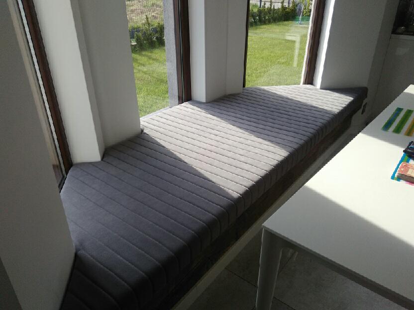 Szary, prążkowany materac na siedzisku wnęki okiennej, z widokiem na ogród i fragmentem białego stołu obok.